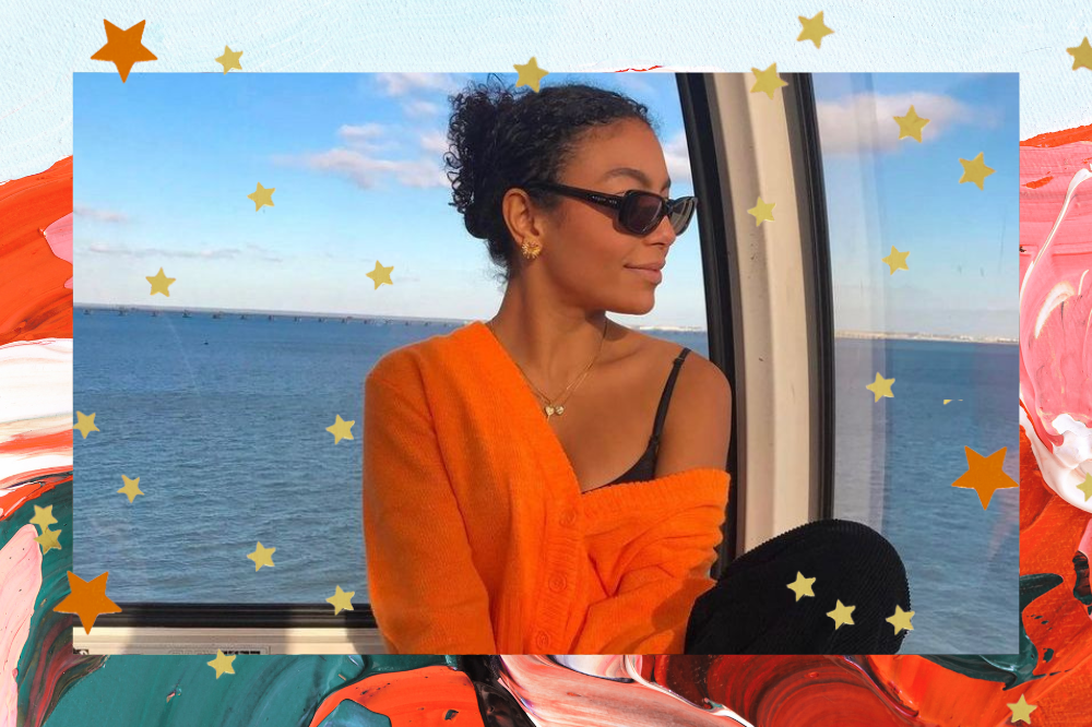 Montagem em fundo com textura de tintas em vermelho, laranja, rosa, branco e verde e estrelinhas douradas espalhadas de foto da Any Gabrielly usando óculos escuro, blusa de manga longa laranja e calça de moletom preta. Ela está sentada, observando a vista de Portugal.