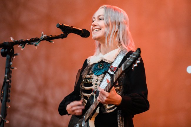 Primavera Sound Festival In Sao Paulo Phoebe Bridgers no palco do Primavera Sound em São Paulo; ela está tocando guitarra enquanto sorri e canta para o público do festival e é iluminada por uma luz alaranjada