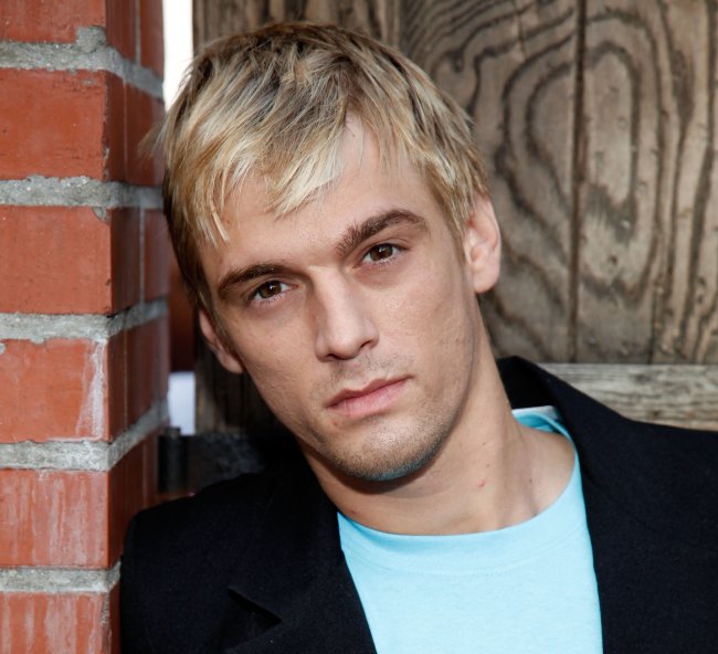 Foto de Aaron Carter encostado em uma parede de tijolos. Ele é um homem branca, loiro e magro