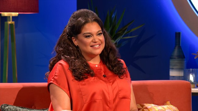 THE CIRCLE (2022) Michelle Buteau, apresentadora do The Circle US, sentada e sorrindo; ela usa uma blusa alaranjada e está sentada em um sofá com uma parede azul e uma planta ao fundo