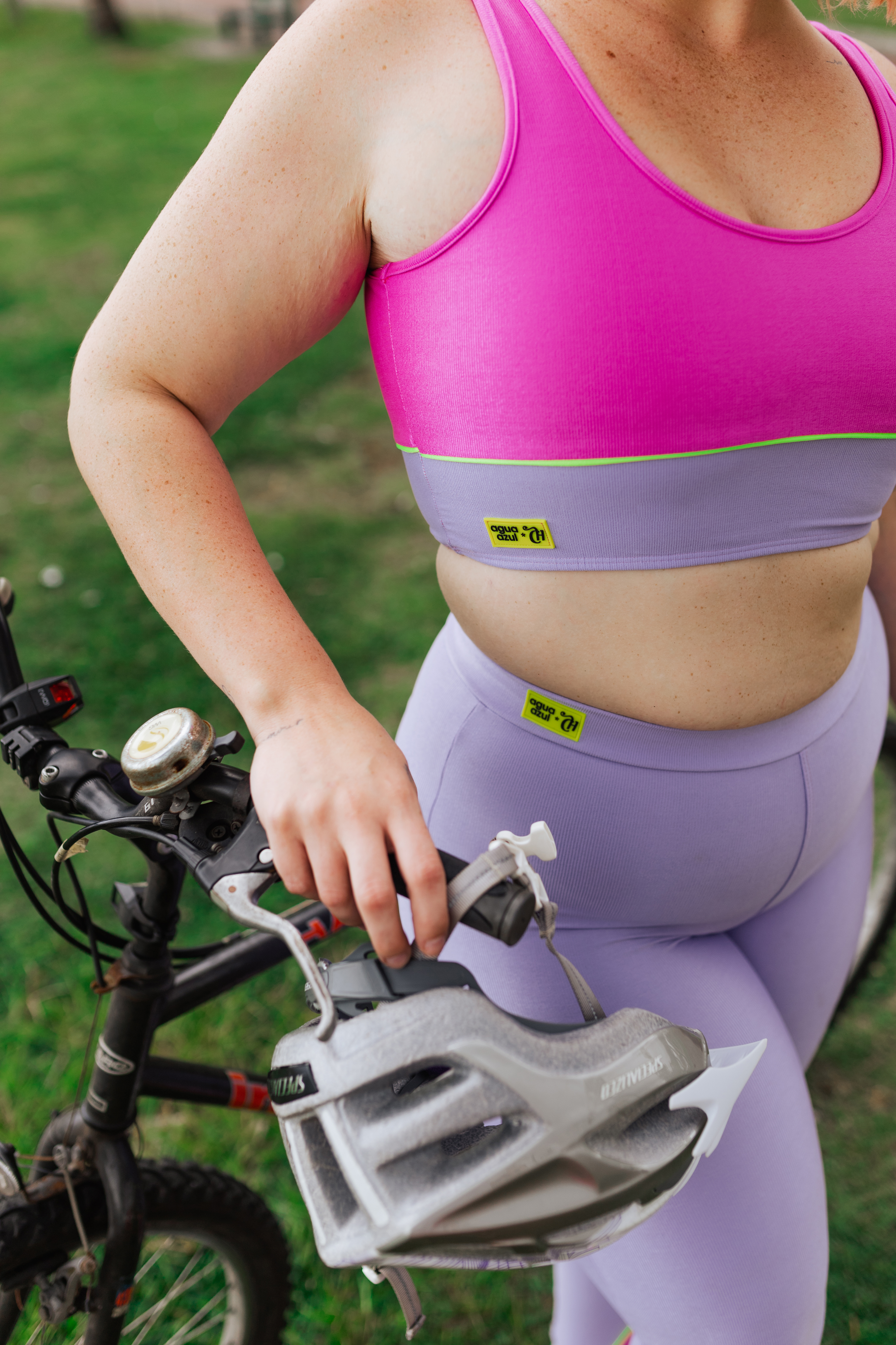 Modelo usando top e legging lilás e rosa da Coleção de roupas da CAPRICHO com a Agua Azul no mood Aguete Welfare ao lado de uma bicicleta em parque