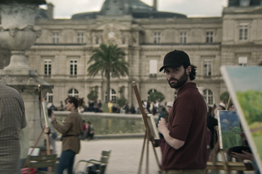 Penn Badgley como Joe Goldberg em Você; ele está em um ambiente externo durante o dia enquanto usa boné preto e observa algo