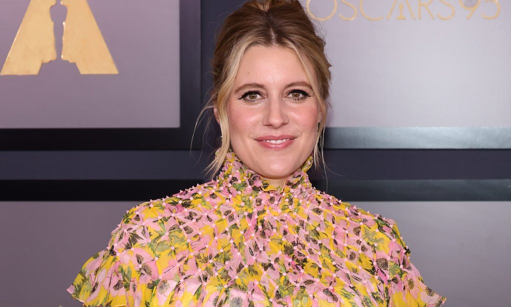 Greta Gerwig posando para foto sorrindo no tapete vemelho do Oscar; ela usa um vestido florido e está com o cabelo preso