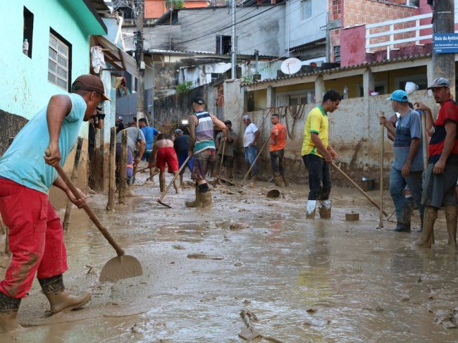 Moradores retiram lama no bairro Itatinga, conhecido como Topolândia, no litoral norte de São Paulo Pessoas com bota e enxada retirando lama