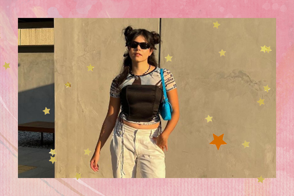 Montagem em fundo rosa com estrelinhas douradas de foto de garota usando óculos de sol, corset preto por cima de camiseta, bolsa azul pendurada nos ombros e calça bege