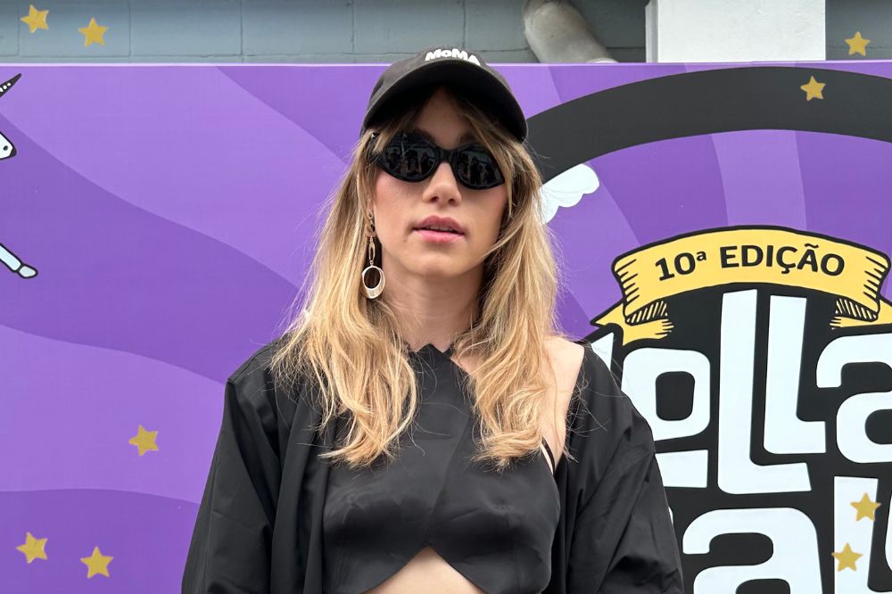 Suki Waterhouse posando para foto com expressão neutra; ela usa óculos escuros e boné preto; ao fundo um backdrop na cor roxa com o logo da 10ª edição do Lollapalooza BR; estrelas amarelas decoram a imagem