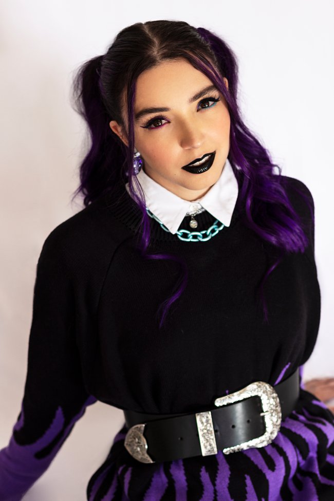 Ana Xisdê encara a cêmera enquanto usa batom preto e delineado. Seu cabelo roxo está preso em maria chiquinha e veste blusa de manga comprida preta com gola branca aparente, cinto grande com fvela prata e saia preta também.