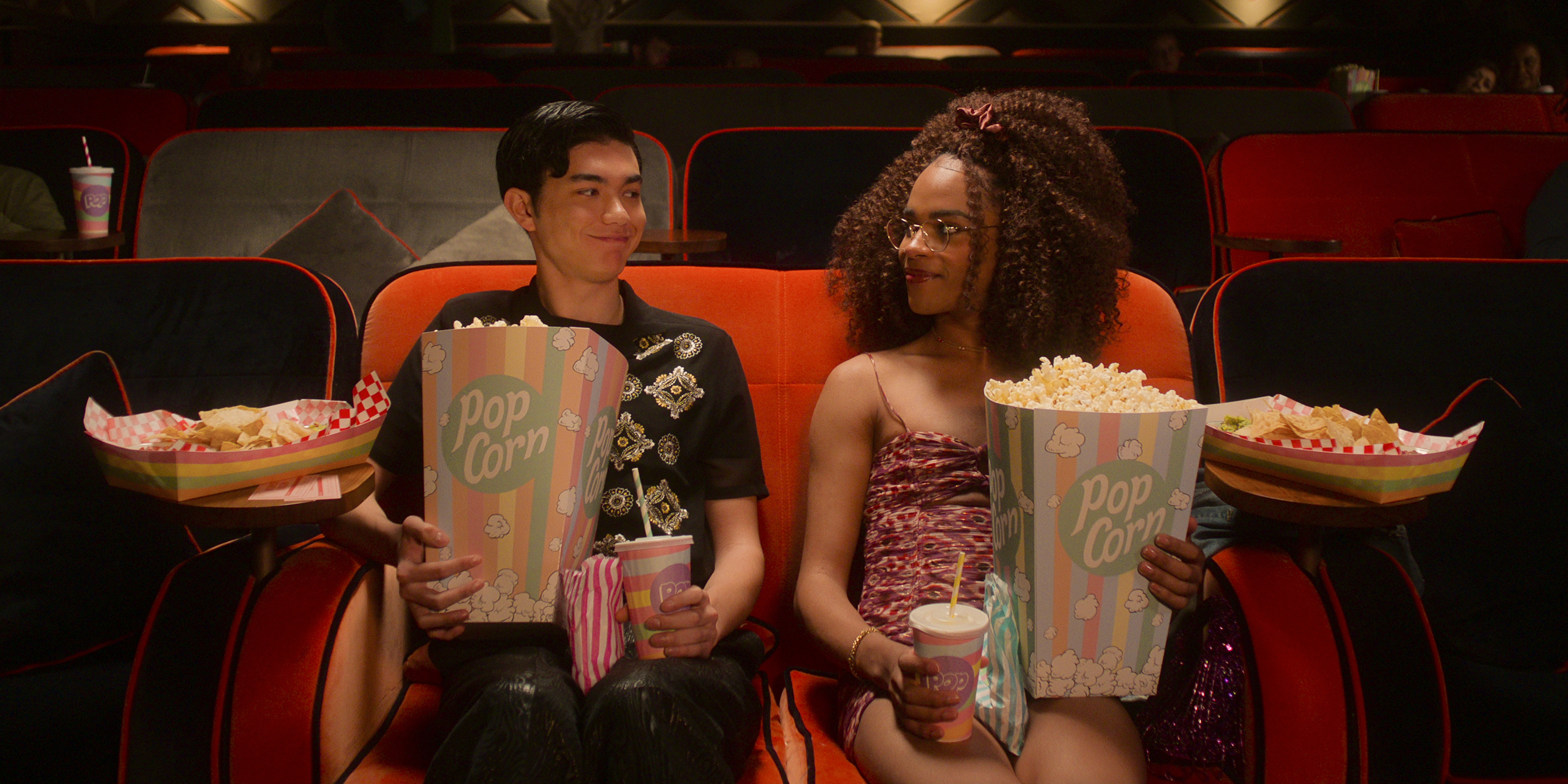 Heartstopper Season 2 Will Gao e Yasmin Finney em cena de Heartstopper; eles estão no cinema se olhando enquanto seguram grandes pacotes de pipoca e bebidas; eles sorriem levemente um para o outro