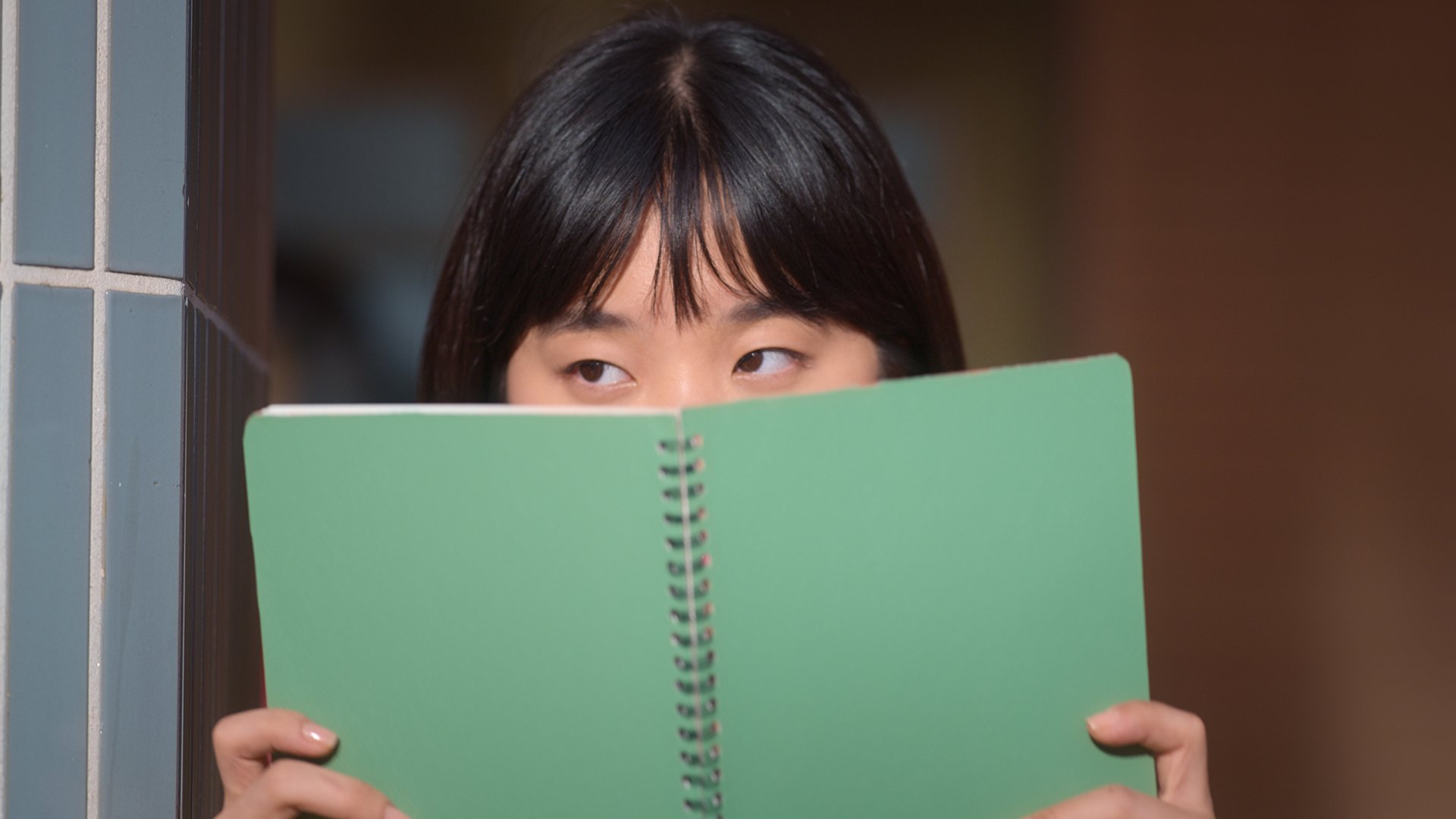 Sharon Cho em cena de Além do guarda-roupa; ela está com um caderno verde escondendo parte do rosto