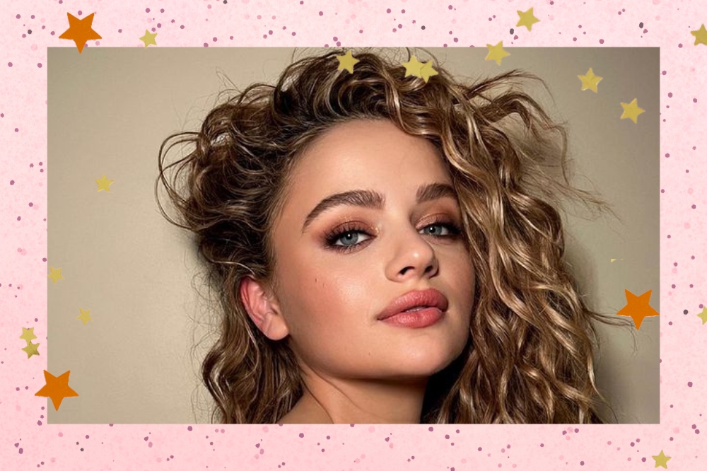 Montagem com fundo rosa. Imagem de Joey King no centro, foco no rosto, o cabelo está jogado para o lado. Ela usa uma maquiagem de festa.