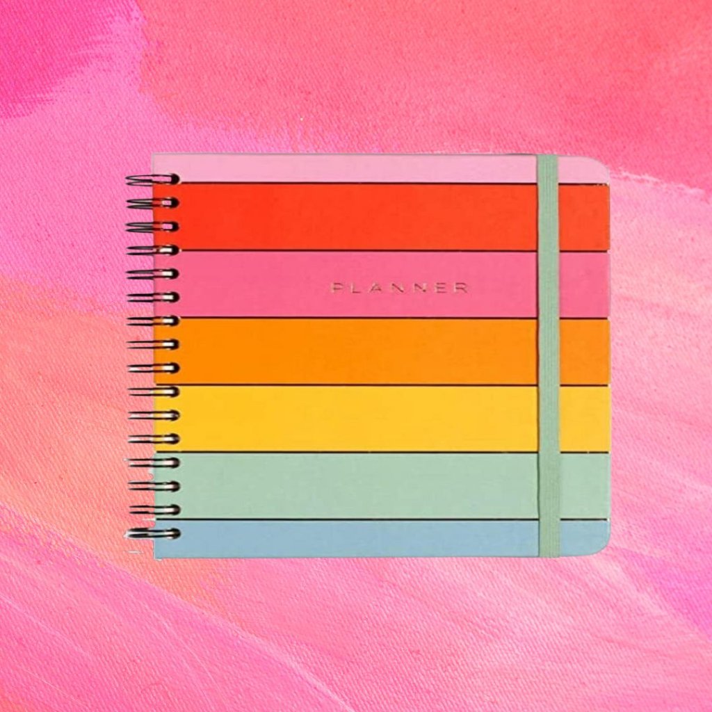 Planner com listras em diferentes cores formando um arco-íris; o fundo é uma textura em tons de rosa e branco