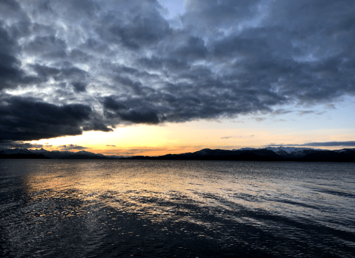 Na imagem, aparece o por do sol amarelado refletindo no mar e em cima, nuvens cinxentas cobrem o cel