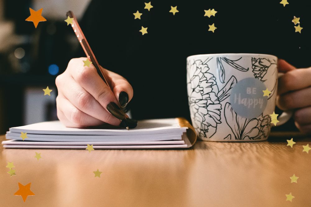A mão de uma mulher, com unas longas e pintadas de preto, escrevem em um caderno. Ao lado do caderno na mesa tem uma xícara