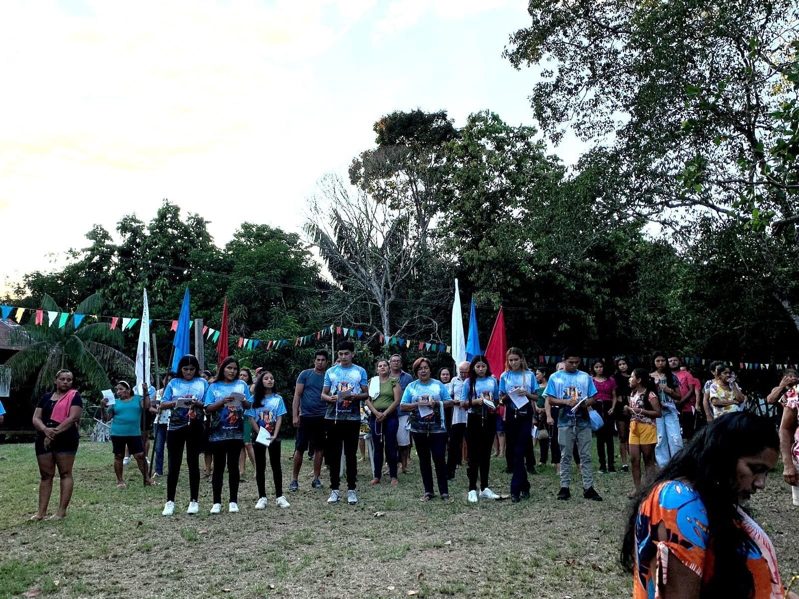 festa-tumbira Em um gramado e árvores de fundo, dezenas de pessoas estão em pé. Na frente, jovens