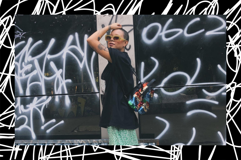 Garota usando camiseta preta, saia midi verde, oxford com meia branca e mochila de paetês nas costas