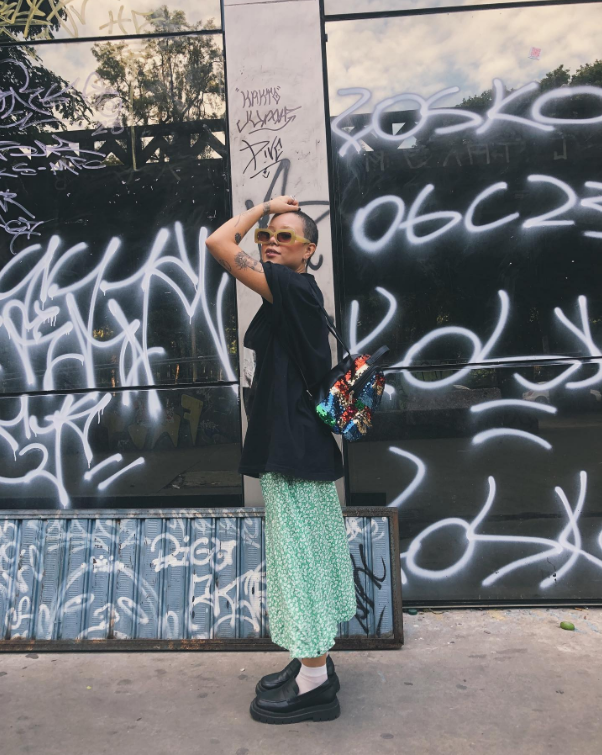 Garota usando camiseta preta, saia midi verde, oxford com meia branca e mochila de paetês nas costas