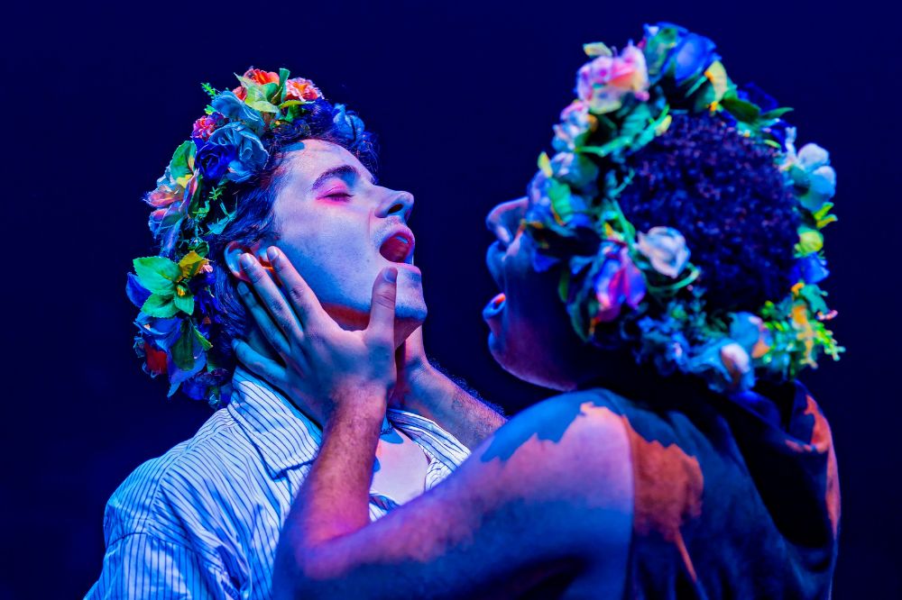 Foto de dois homens cantando de frente um para o outro, usando uma coroa de flores. Um deles segura o rosto do outro, iluminação azul.