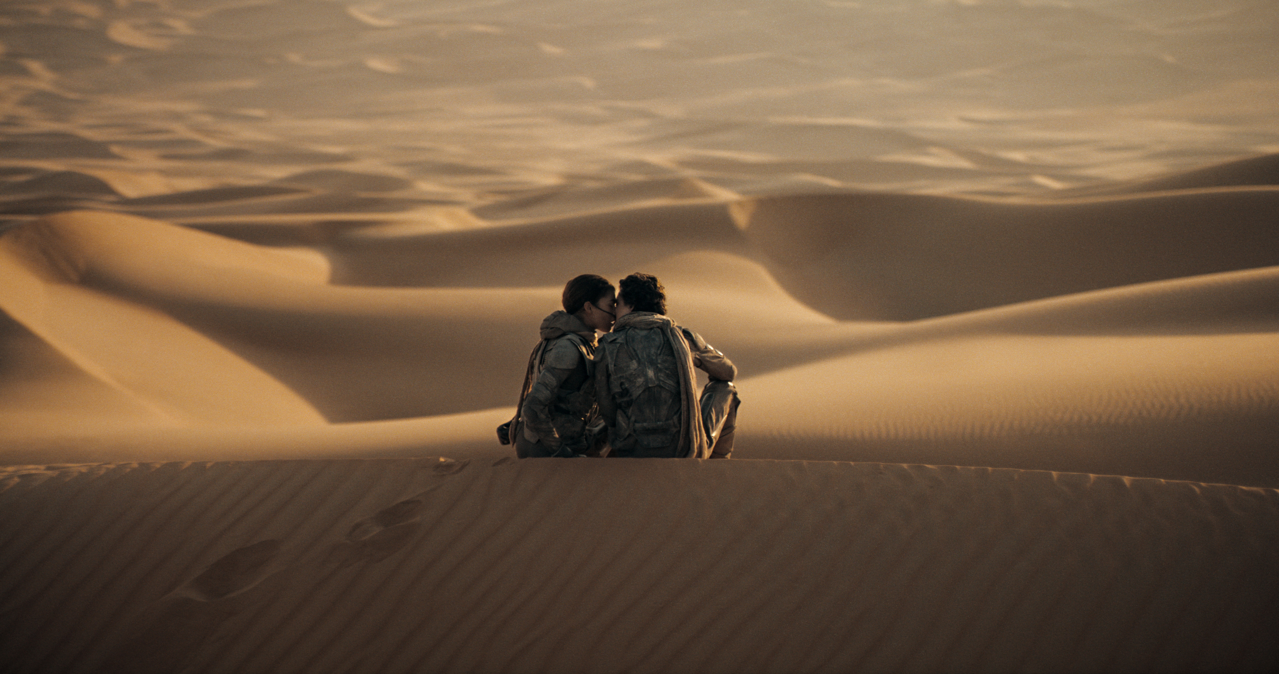 rev-1-DUN2-T3-0026r_High_Res_JPEG Zendaya e Timotheé Chalamet como Chani e Paul em cena de Duna Parte 2; eles estão no deserto sentados nas dunas de areia enquanto se beijam