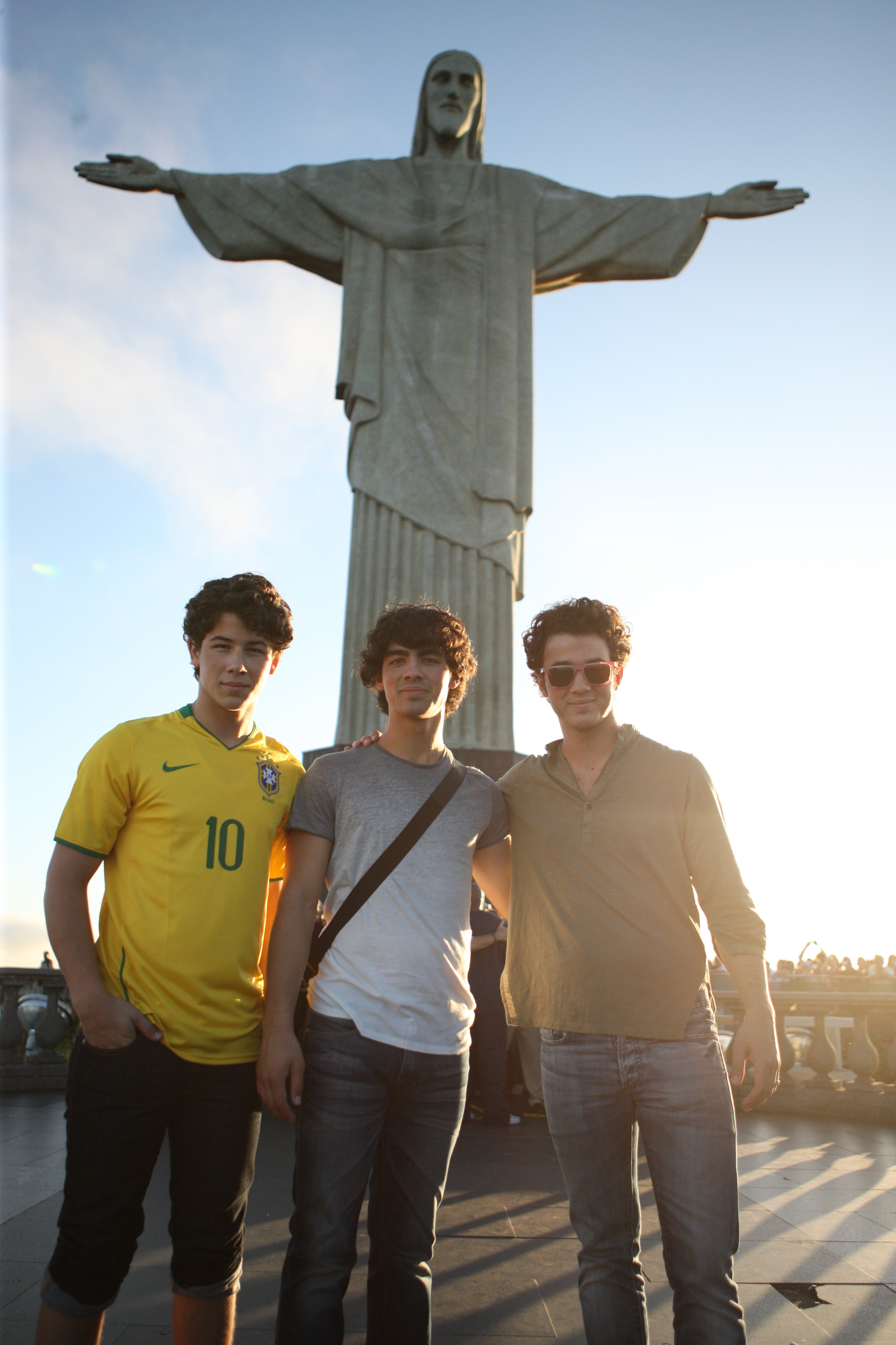 The Jonas Brothers Visit Christ The Redeemer Statue In Rio De Janeiro Nick, Joe e Kevin Jonas, dos Jonas Brothers, visitando o Cristo Redentor no Rio de Janeiro; eles estão sorrindo levemente e posando para foto na frente da estátua durante o dia
