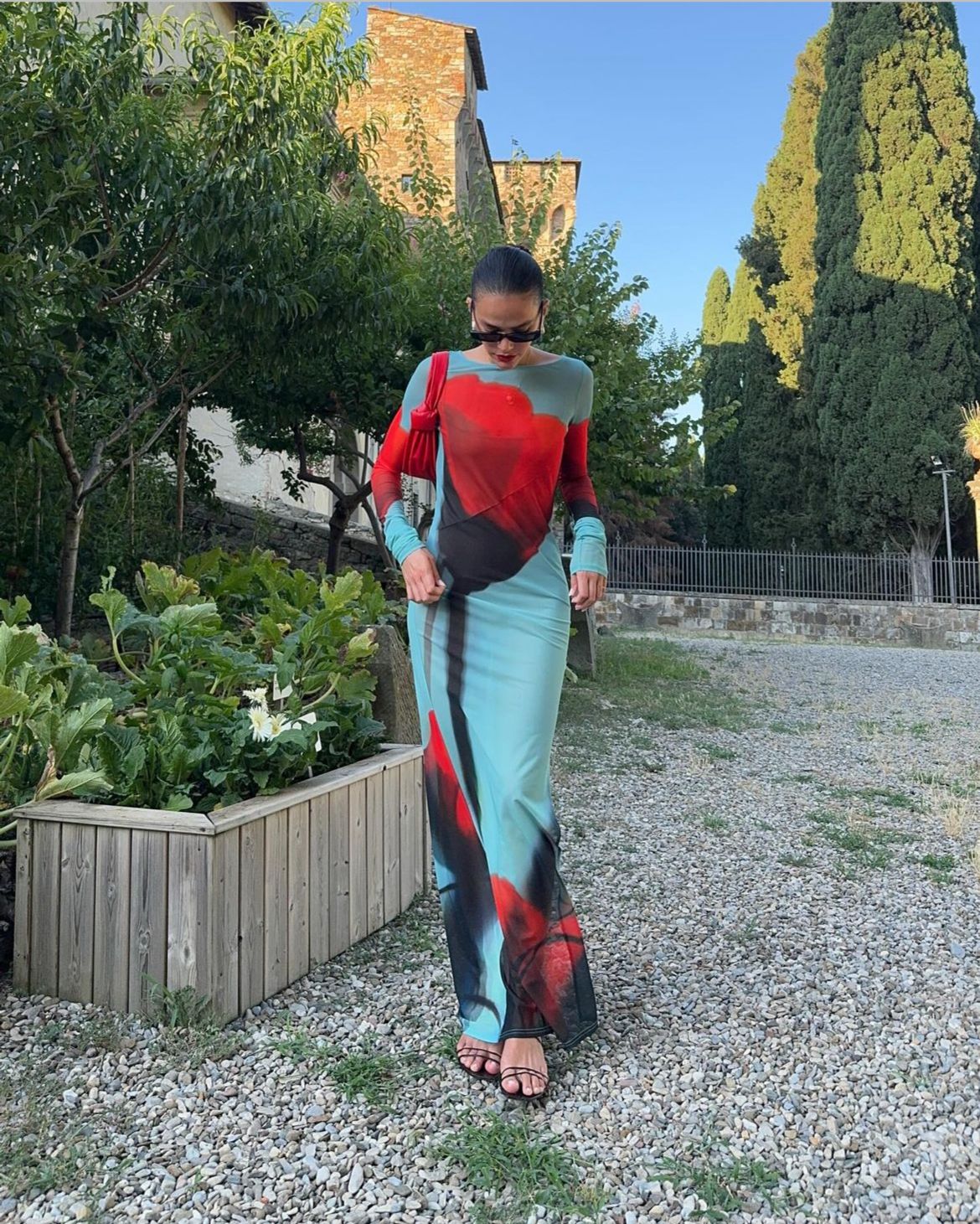 Look Bruna Marquezine transparente com os pés de fora Imagem de corpo inteiro de Bruna Marquezine, mulher branca de cabelos pretos. Ela está olhando para baixo em um lugar com jardim, vestindo um vestido longo justo com transparência vermelho, preto e azul.