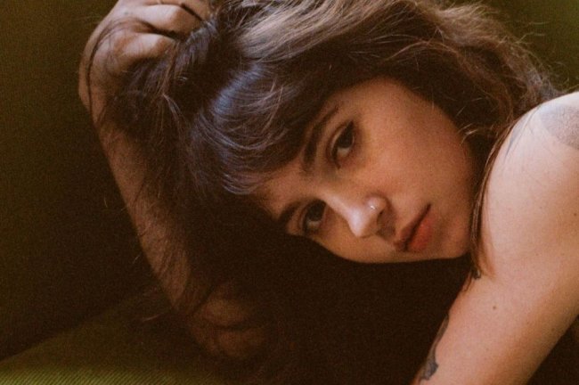 clairo Imagem de Clairo de lado posando para câmera com a cabeça apoiada em um dos braços; ela está com a expressão neutra
