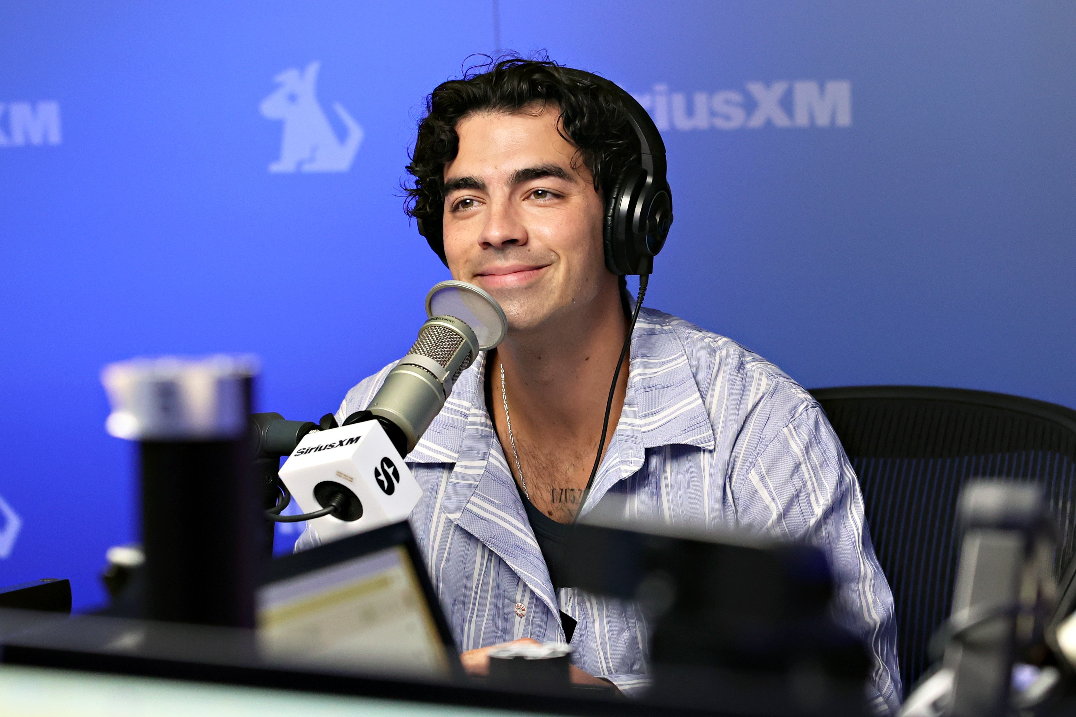 Joe Jonas em entrevista de rádio sorrindo levemente com microfone e fone de ouvido