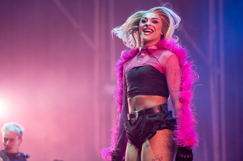 Pabllo Vittar se apresentando enquanto sorri e pula em um palco com iluminação nas cores rosa e azul