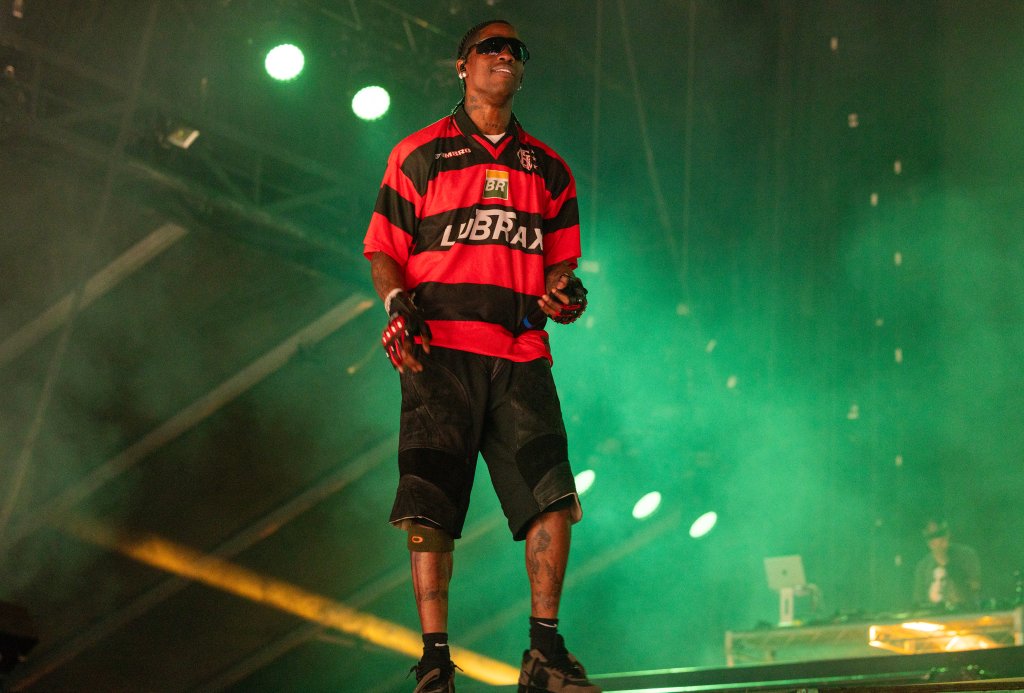 Travis Scott se apresentando com camisa do Flamengo; ele está sorrindo enquanto observa o público e o palco tem iluminação na cor verde