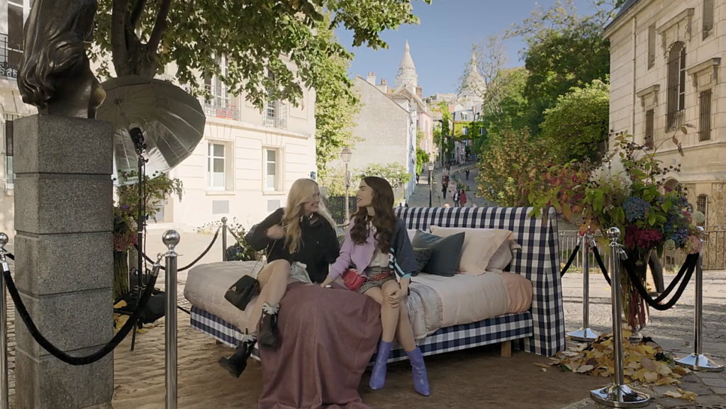 Cena de Emily em Paris, com Emily e Camille sentadas em uma cama no meio de uma praça.