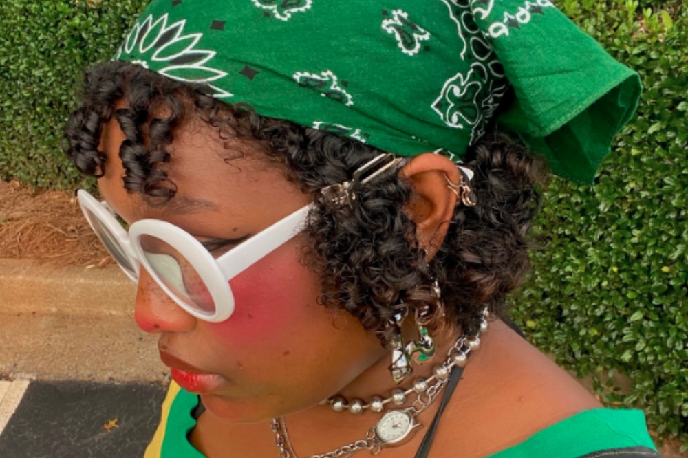 Garota usando bandana verde no cabelo, óculos brancos redondos e colares olhando para o lado com cabeça baixa