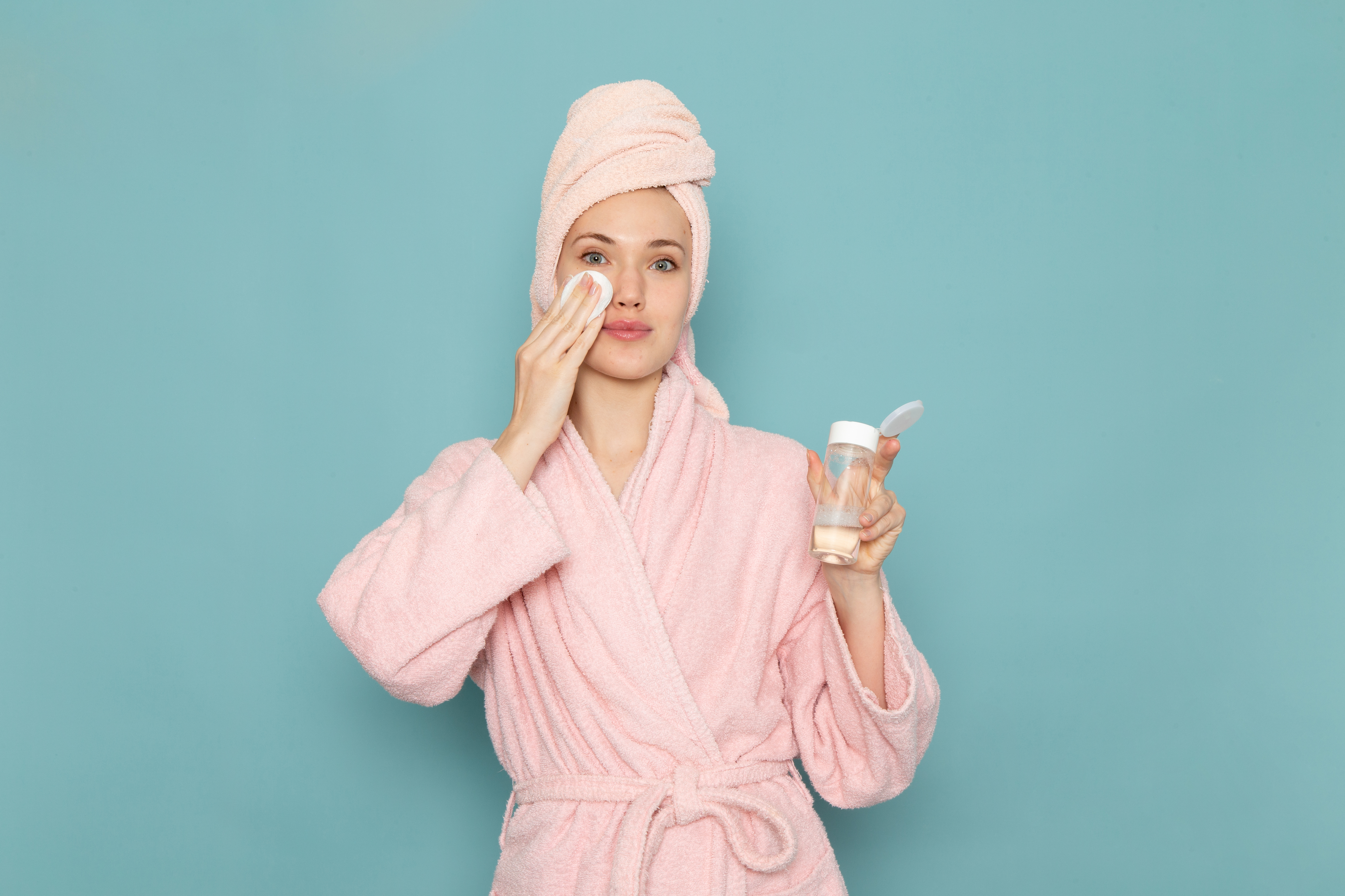 tonico-facial-pele Mulher usando roupão rosa enquanto aplica tônico facial em um algodão