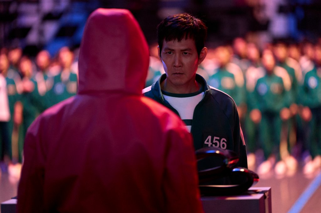 Lee Jung-jae como Seong Gi-hun na 2ª temporada de Round 6; ele está com expressão séria enquanto olha para um dos seguranças mascarados do jogo, que aparece de costas com o macacão vermelho