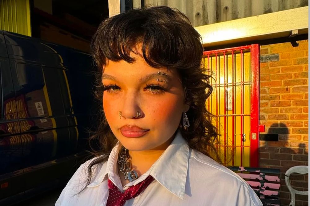 Foto da cantora Lola Young. Ela possui franja e cabelo curto vermelho, veste uma camisa branca e gravata vermelha.
