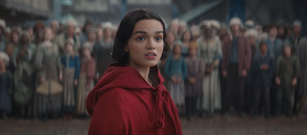 Rachel Zegler como Branca de Neve em live-action da Disney; ela está em cena usando uma capa vermelha com o povo do reino ao fundo