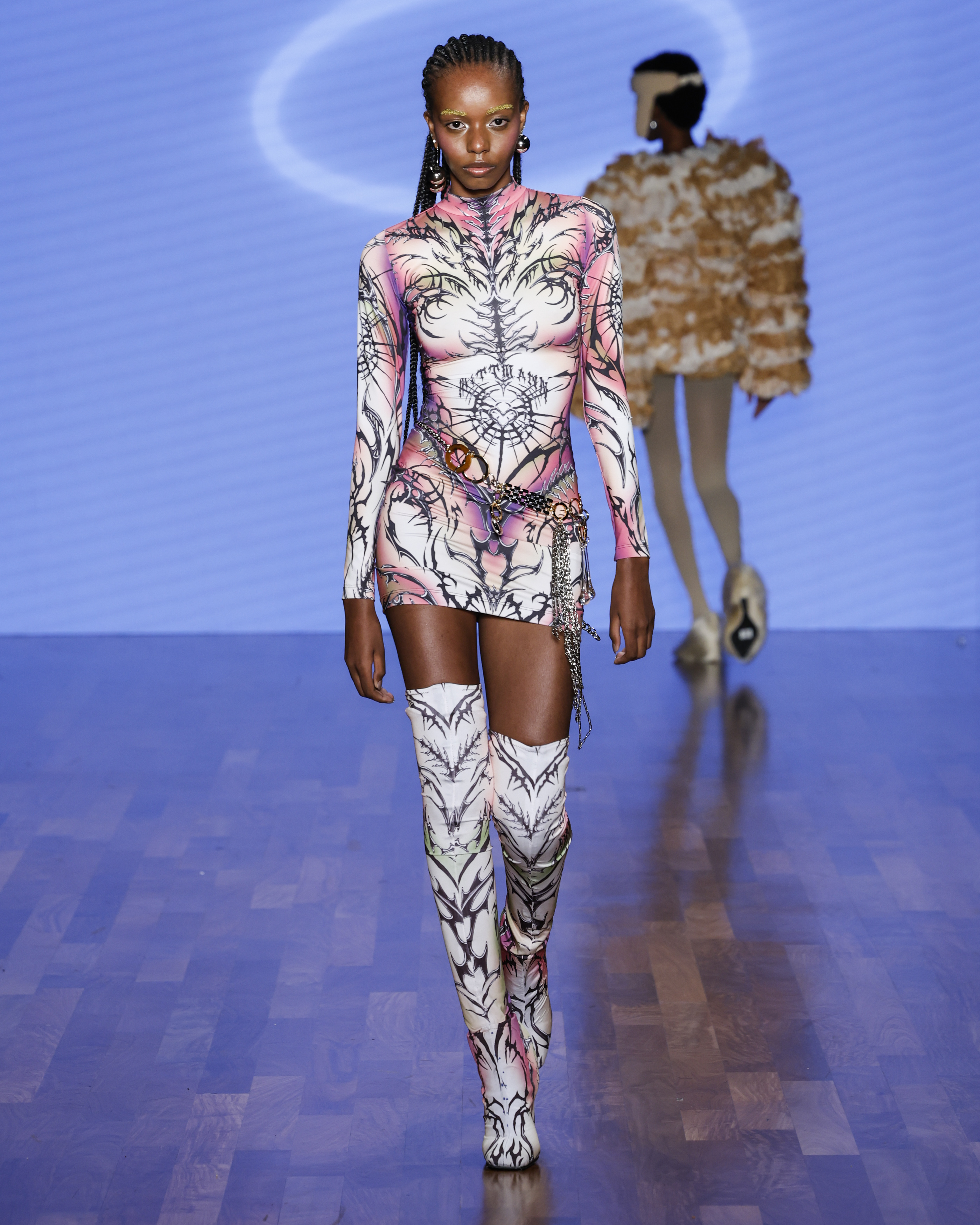 Dario MittmannSPFW N59Foto: Ze Takahashi/ @agfotosite Modelo desfile na passarela de Dário Mittmann com um vestido curto de manga longa em conjunto com botas altas. A estampa das peças possui desenhos que remetem ao cybersigilism.