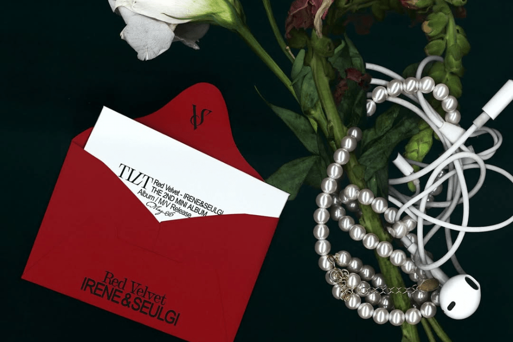 Envelope vermelho com o nome de IRENE & SEULGI, rosas brancas, colar de perolas e fones de ouvido