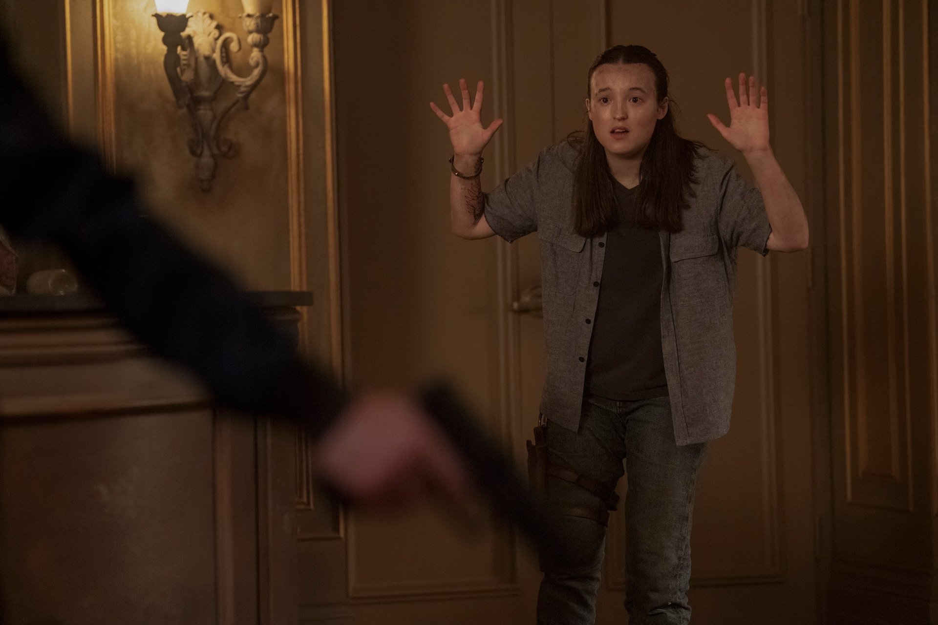 Bella Ramsey como Ellie em cena de The Last of Us; ela está com os braços levantados enquanto Abby segura uma arma que aparece na imagem fora de foco