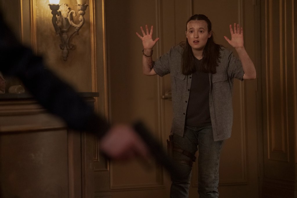 Bella Ramsey como Ellie em cena de The Last of Us; ela está com os braços levantados enquanto Abby segura uma arma que aparece na imagem fora de foco