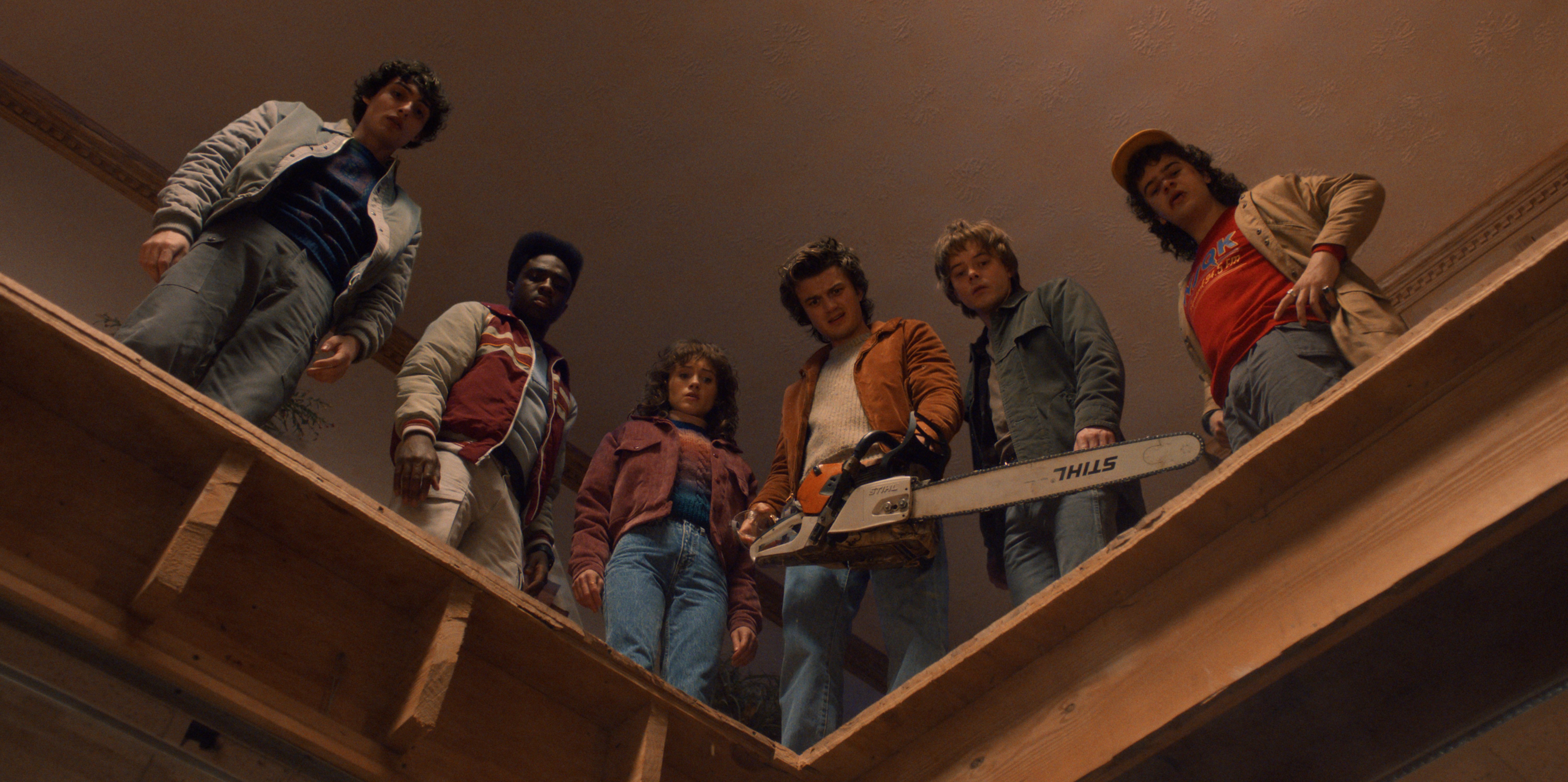Finn Wolfhard como Mike Wheeler, Caleb McLaughlin como Lucas Sinclair, Natalia Dyer como Nancy Wheeler, Joe Keery como Steve Harrington, Charlie Heaton como Jonathan Byers, e Gaten Matarazzo como Dustin Henderson em cena de Stranger Things; eles estão reunidos olhando para baixo