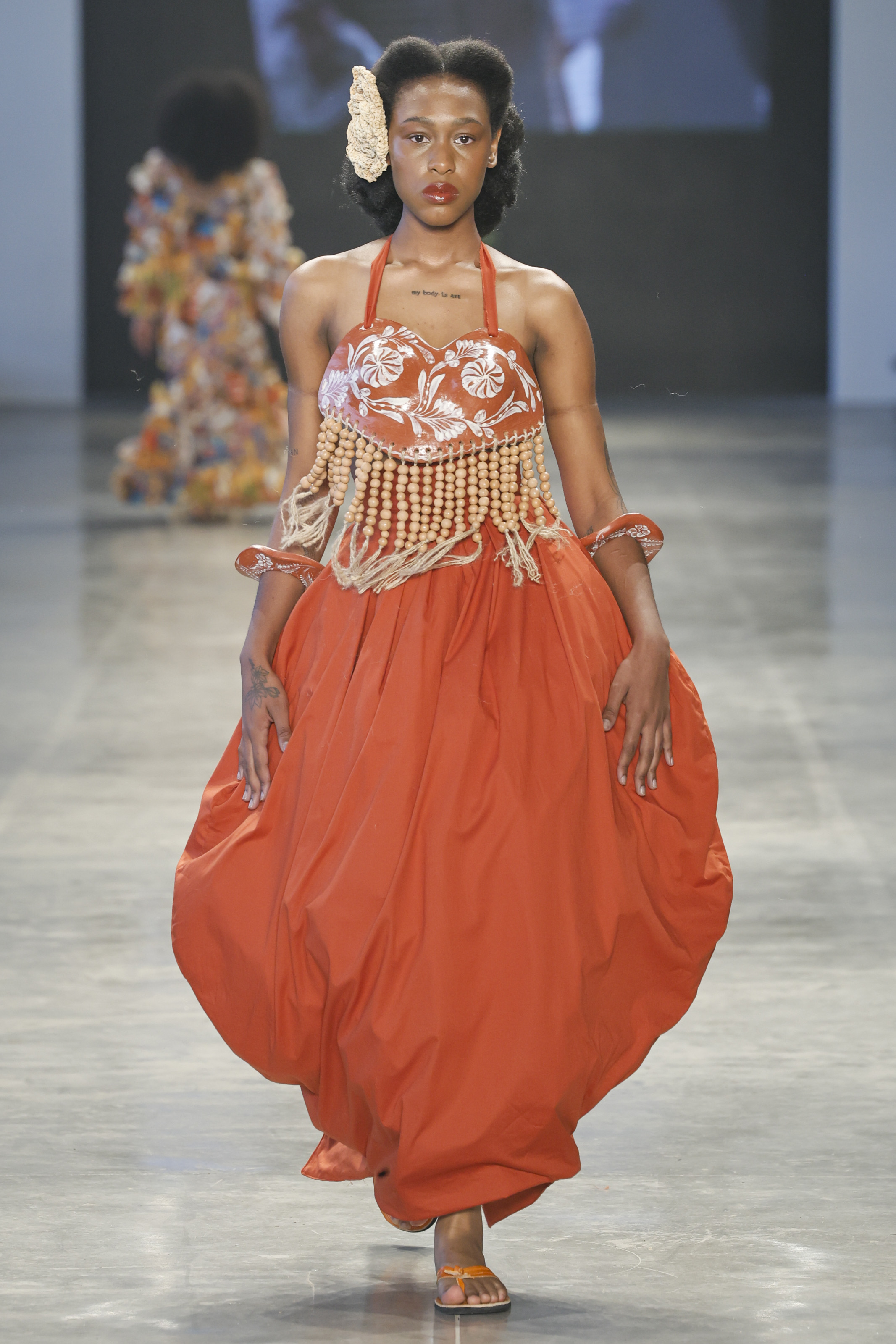 Modelo desfilando no desfile da marca Meninos Rei SPFW N60 com look laranja, pele iluminada e lábios avermelhados