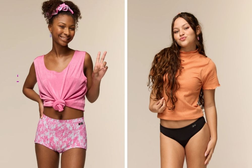 Duas jovens posam em um estúdio com fundo neutro. À esquerda, uma garota usa um top rosa amarrado e uma calcinha estampada em tons de rosa, fazendo o sinal de paz e sorrindo. À direita, outra garota veste uma blusa laranja e uma calcinha preta, fazendo biquinho e gesto de paz com a mão. Ambas aparentam estar confortáveis e confiantes.