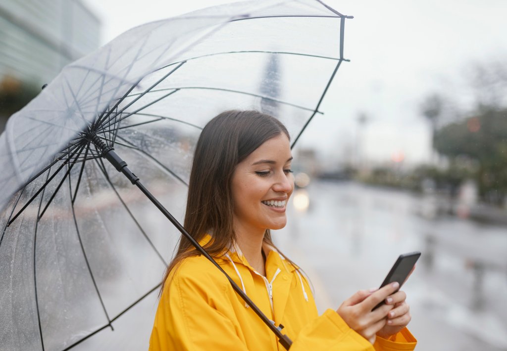 Mulher segurando guarda-chuva enquanto olha o celular e usa capa de chuva amarela