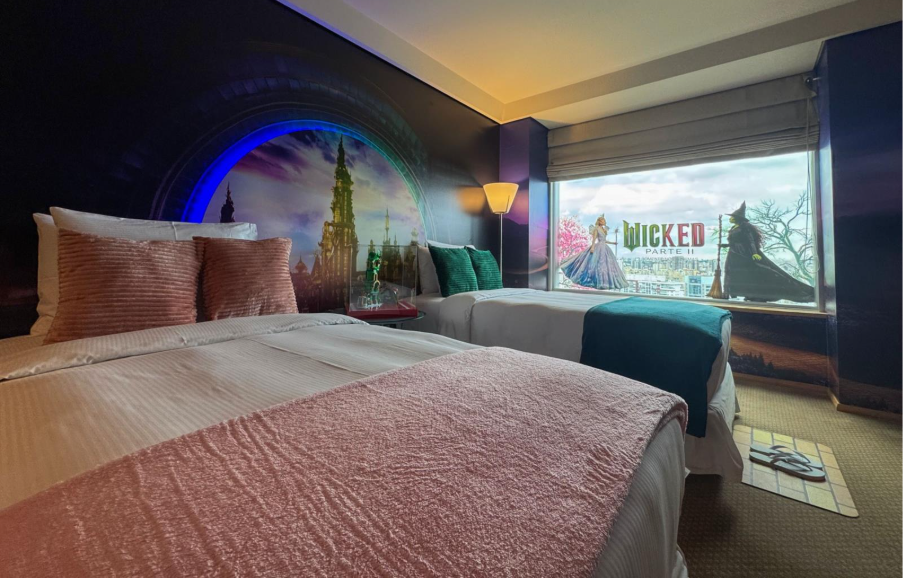 Quarto de hotel decorado com o tema do filme Wicked: Parte II. O ambiente tem duas camas — uma com roupa de cama rosa e outra com verde — e uma parede ilustrada com um castelo iluminado por luzes mágicas. Na janela, há uma imagem dos personagens principais de Wicked, com o logotipo do filme ao centro. O quarto tem iluminação suave e vista para a cidade ao fundo.