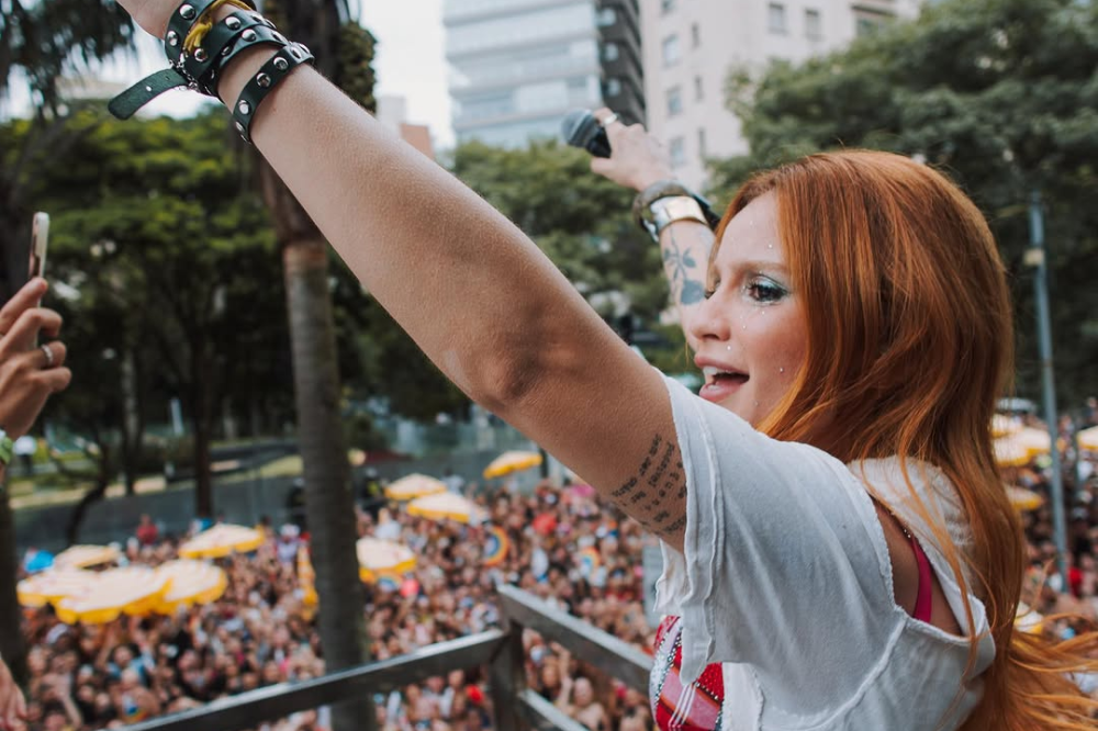 Cantora ruiva em trio elétrico levanta o braço e segura um microfone enquanto se apresenta para uma multidão em avenida cercada por prédios e árvores, durante evento ao ar livre em clima de festa.