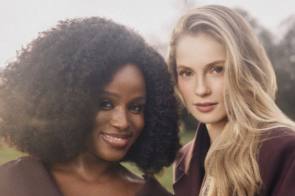 Duas mulheres posam lado a lado ao ar livre: à esquerda, uma mulher negra com cabelo crespo volumoso sorri para a câmera; à direita, uma mulher loira de cabelo longo também encara a câmera com expressão suave, ambas usando roupas em tons neutros.