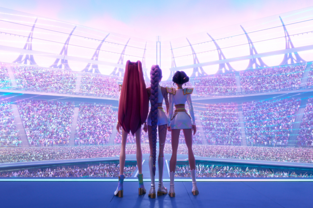 Três personagens animadas vistas de costas estão em um palco futurista, olhando para um enorme estádio lotado de público. Elas usam figurinos brilhantes e coordenados, com botas de salto e detalhes metálicos, enquanto luzes coloridas iluminam a cena em tons pastel, criando um clima de show grandioso e pop.