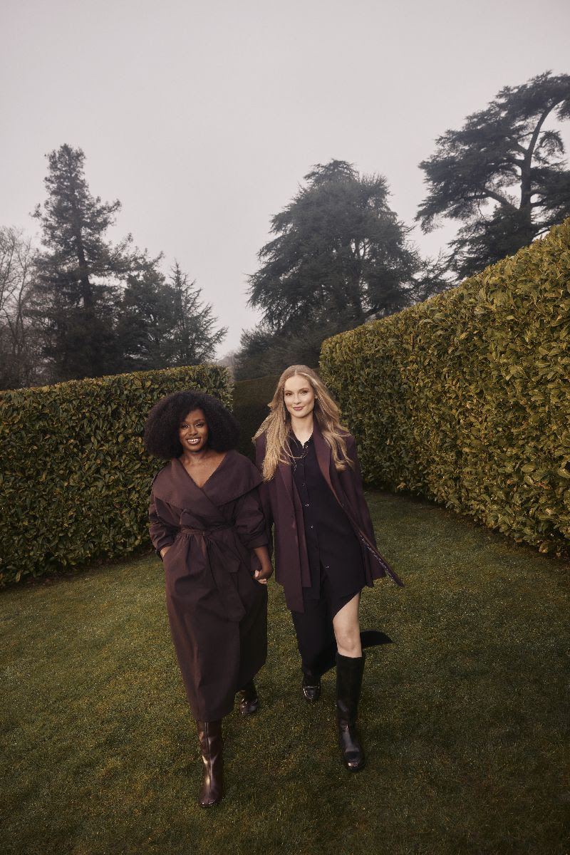 Duas mulheres caminham juntas em um jardim cercado por sebes altas: à esquerda, uma mulher negra com cabelo crespo usa vestido marrom e sorri; à direita, uma mulher loira de cabelo longo veste look escuro com botas, enquanto ambas seguem em direção à câmera em um ambiente ao ar livre com árvores ao fundo.
