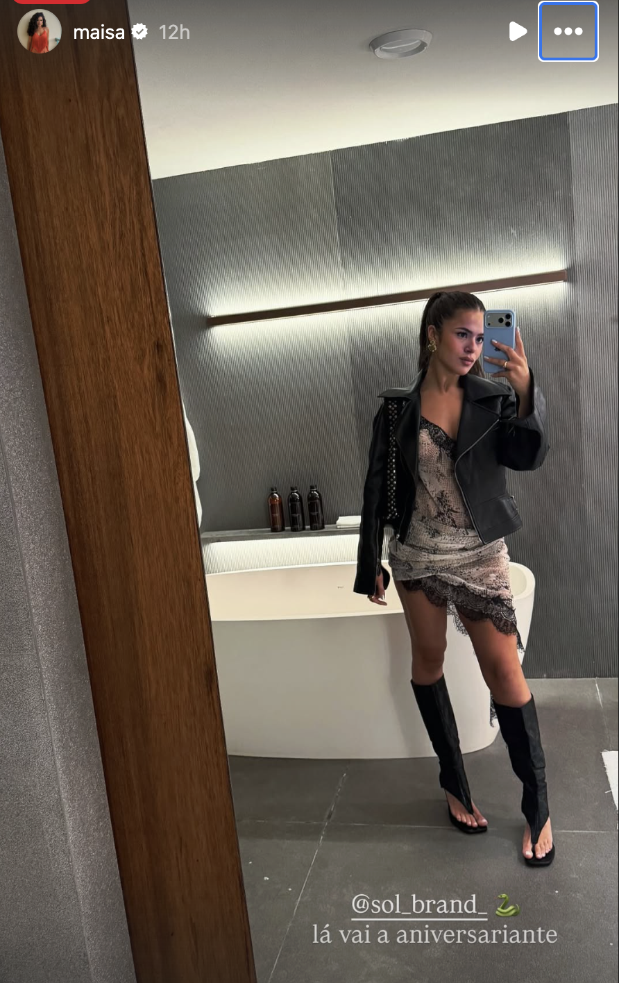 Maisa Silva, com rabo de cavalo e maquiagem leve, tira selfie no espelho de um banheiro moderno. Ela veste um vestido curto de renda floral, jaqueta de couro preta com tachas e botas pretas de salto alto com abertura nos dedos. Ao fundo, uma banheira branca e parede cinza com iluminação embutida