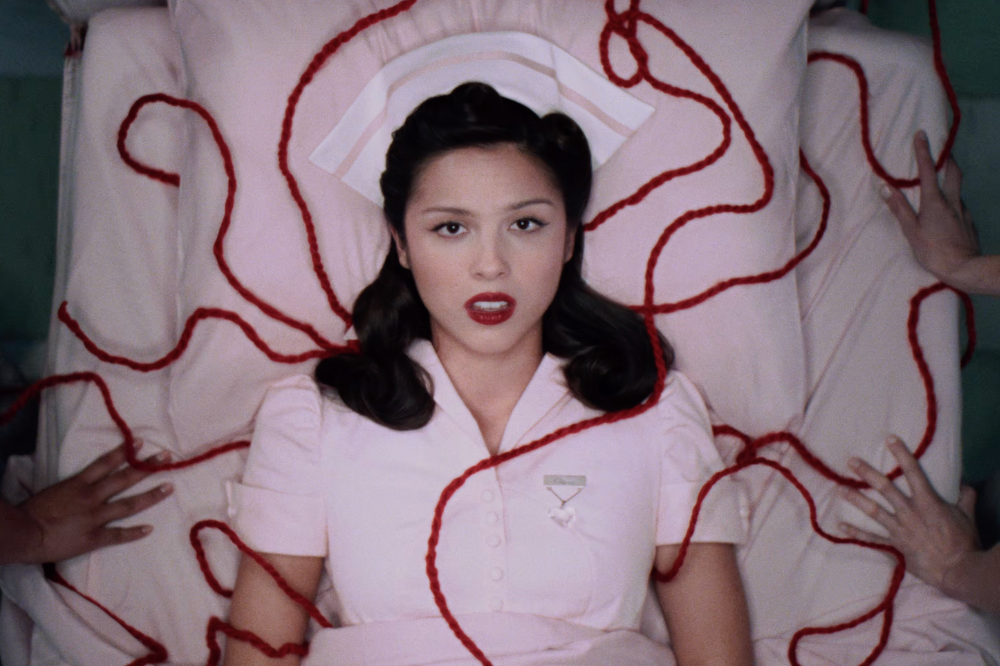 Cantora Olivia Rodrigo deitada em uma cama, vestindo uniforme de enfermeira branco e touca, com lábios vermelhos. Fios de lã vermelhos se espalham sobre ela e a cama, enquanto mãos de diferentes tons de pele tocam a lã e o lençol ao redor