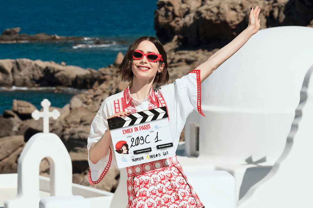 Atriz Lily Collins sorrindo, usando óculos de sol vermelhos, blusa branca com detalhes vermelhos e saia estampada, segurando uma claquete de "Emily in Paris", com o mar e construções brancas ao fundo